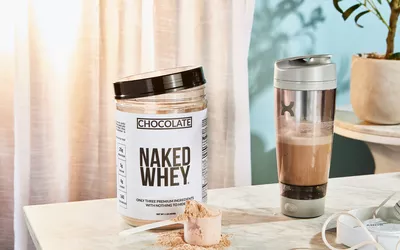 Naked Nutrition Chocolate Whey Protein Powder in a scoop next to its container and a blender bottle