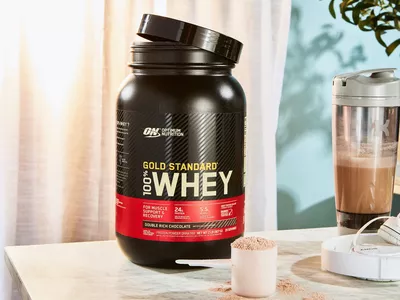 Optimum Nutrition Gold Standard 100% Whey Protein Powder in a scoop next to the jar and blender bottle