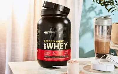 Optimum Nutrition Gold Standard 100% Whey Protein Powder in a scoop next to the jar and blender bottle