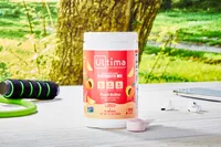 A container of Ultima Replenisher Electrolyte Hydration Powder next to the scoop on a wooden table