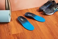 The Walk-Hero Comfort and Support Plantar Fasciitis Insoles on a wood floor next to shoes and exercise equipment. 