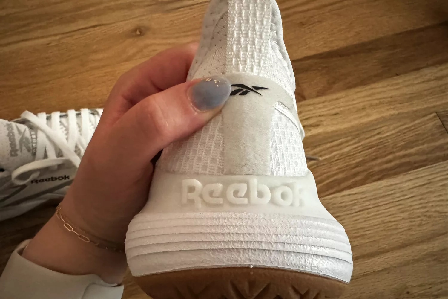 A hand holding the Reebok Nano Court sneaker in the air to show the back with a wooden background