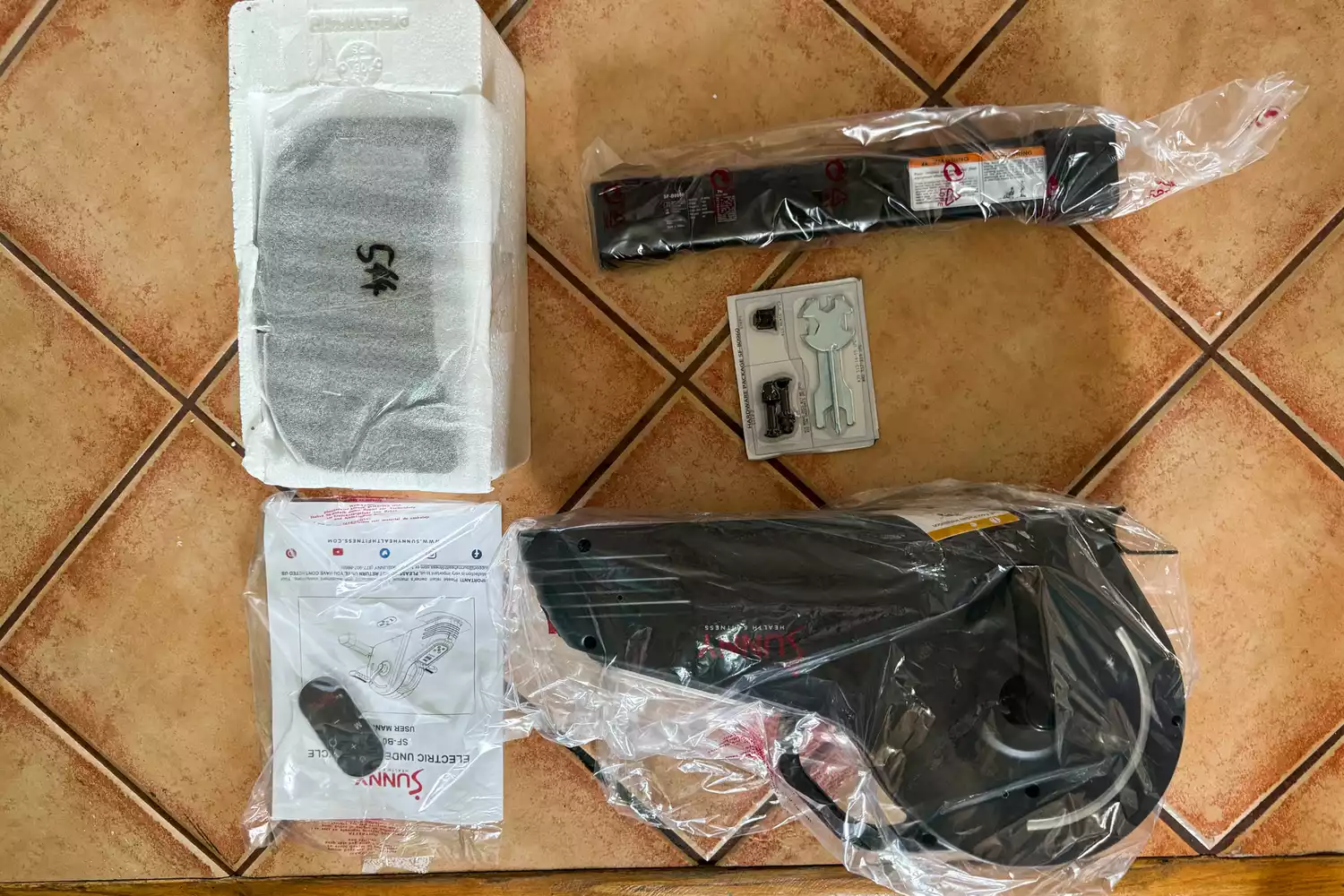 The pieces of the Sunny Health & Fitness SF-B0960 Motorized Electric Under Desk Bike on a tile floor