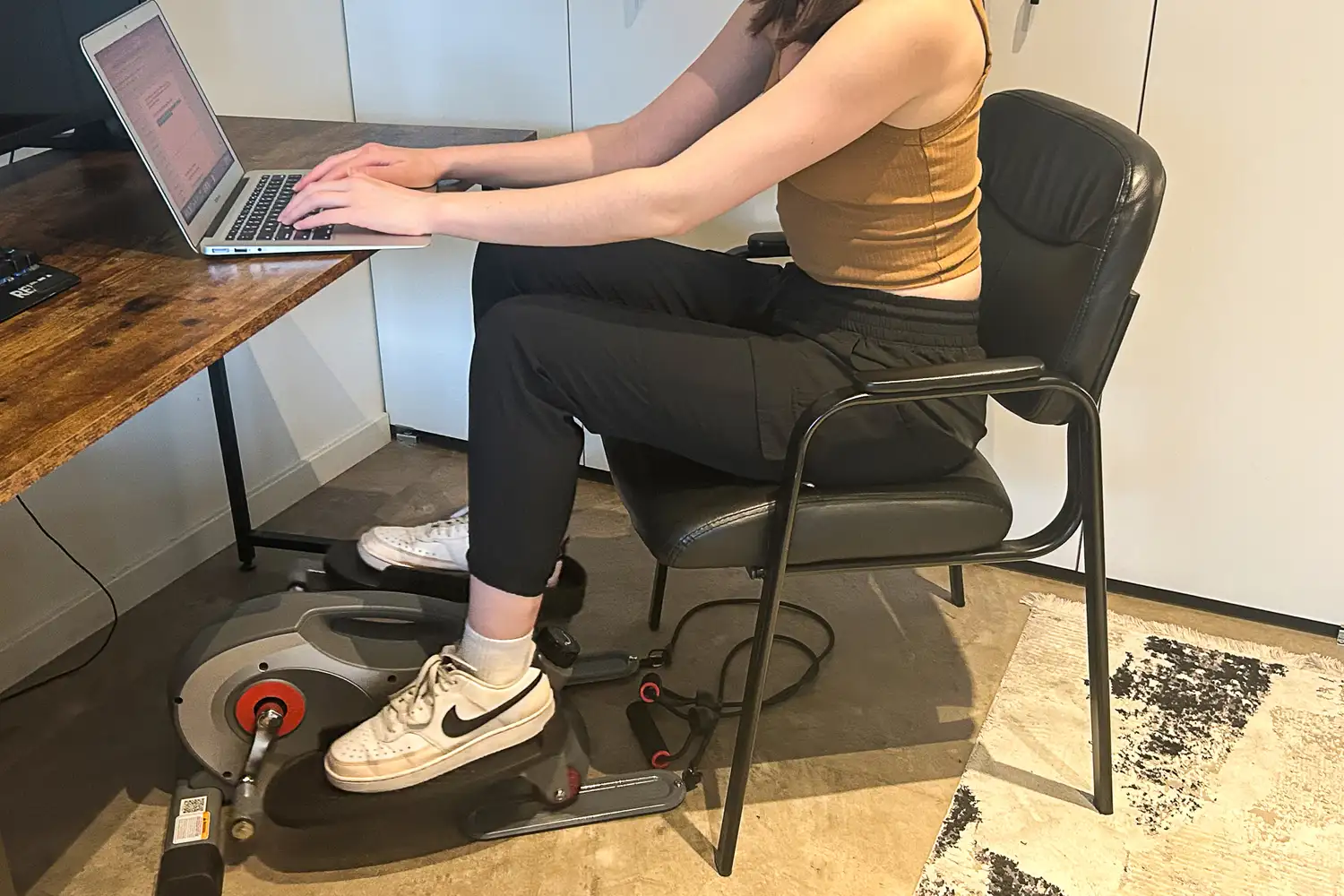 A person uses the Sunny Health & Fitness Smart Stand/Sit Mini Elliptical Exercise Trainer while sitting at a desk