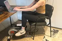 A person uses the Sunny Health & Fitness Smart Stand/Sit Mini Elliptical Exercise Trainer while sitting at a desk