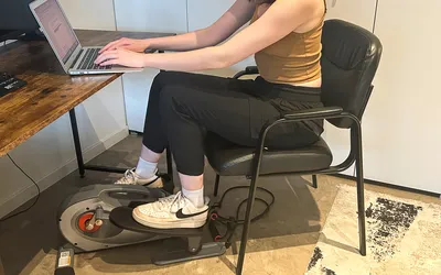 A person sits in a chair while using the Sunny Health & Fitness Smart Stand/Sit Mini Elliptical Exercise Trainer