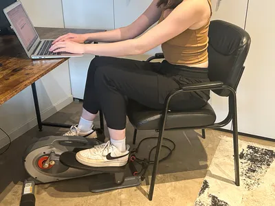 A person sits in a chair while using the Sunny Health & Fitness Smart Stand/Sit Mini Elliptical Exercise Trainer