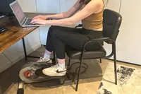 A person sits in a chair while using the Sunny Health & Fitness Smart Stand/Sit Mini Elliptical Exercise Trainer