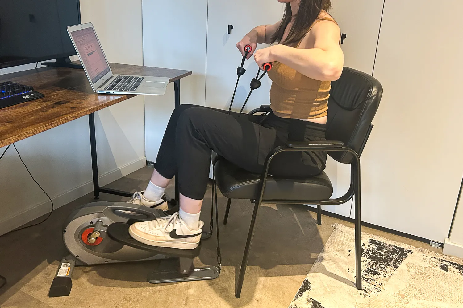 A person uses the Sunny Health & Fitness Smart Stand/Sit Mini Elliptical Exercise Trainer while sitting in a chair