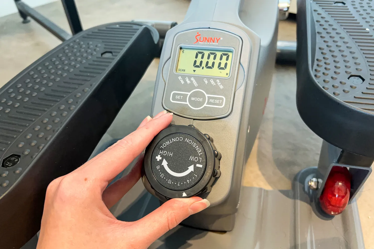 A person turns the tension control dial on the Sunny Health & Fitness Smart Stand/Sit Mini Elliptical Exercise Trainer