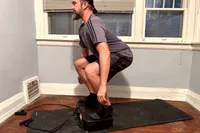 A person squats on the Sunny Health & Fitness Fitboard 2D Vibration Platform With Resistance Band