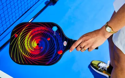 A hand holding a pickleball paddle with a vibrant spiral design on a blue court wearing a watch and rings sneaker and net partially visible