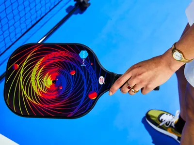 A hand holding a pickleball paddle with a vibrant spiral design on a blue court wearing a watch and rings sneaker and net partially visible