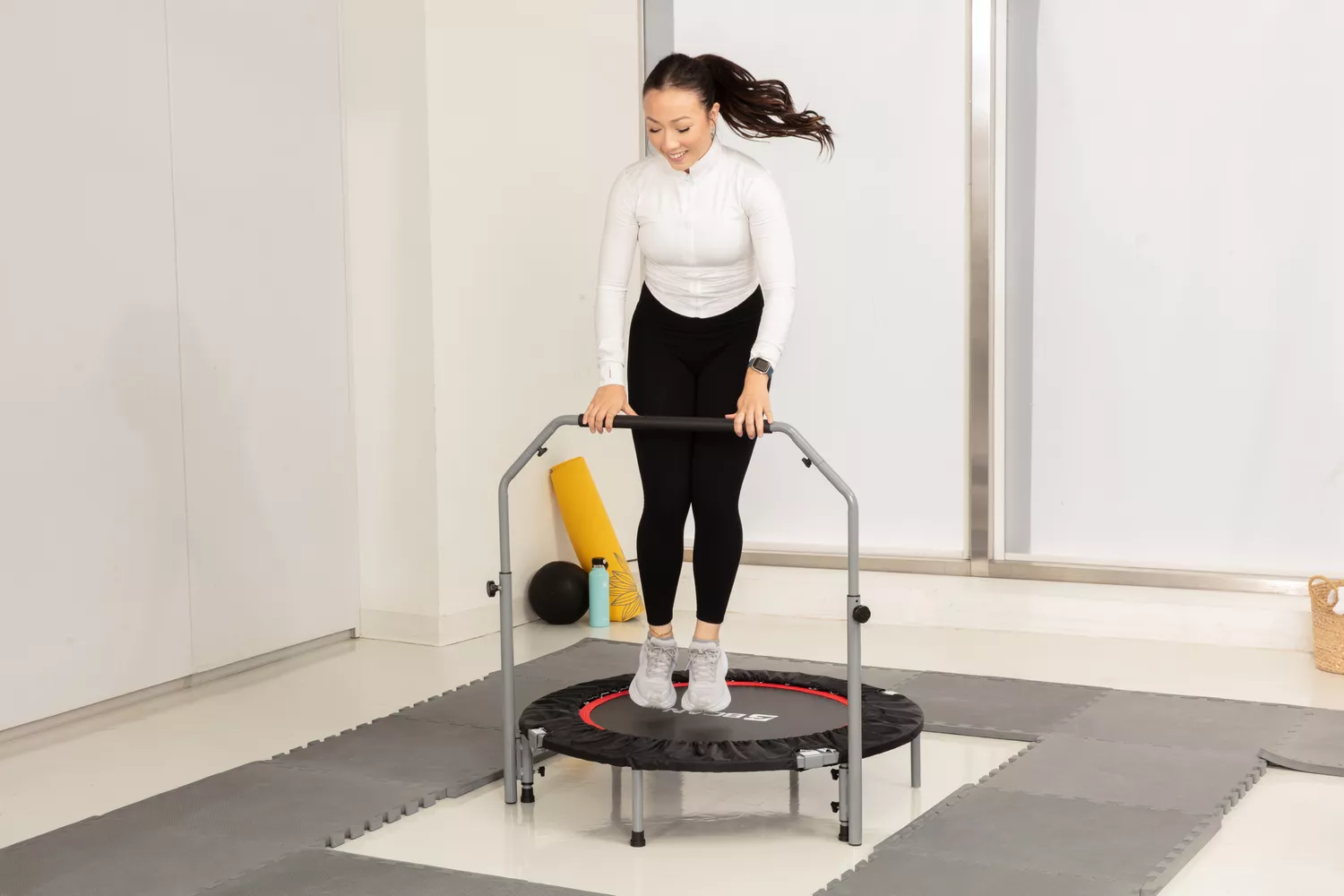 Person holding handle bar while jumping on a BCAN Foldable Mini Trampoline surrounded by padded mats