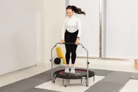 Person holding handle bar while jumping on a BCAN Foldable Mini Trampoline surrounded by padded mats