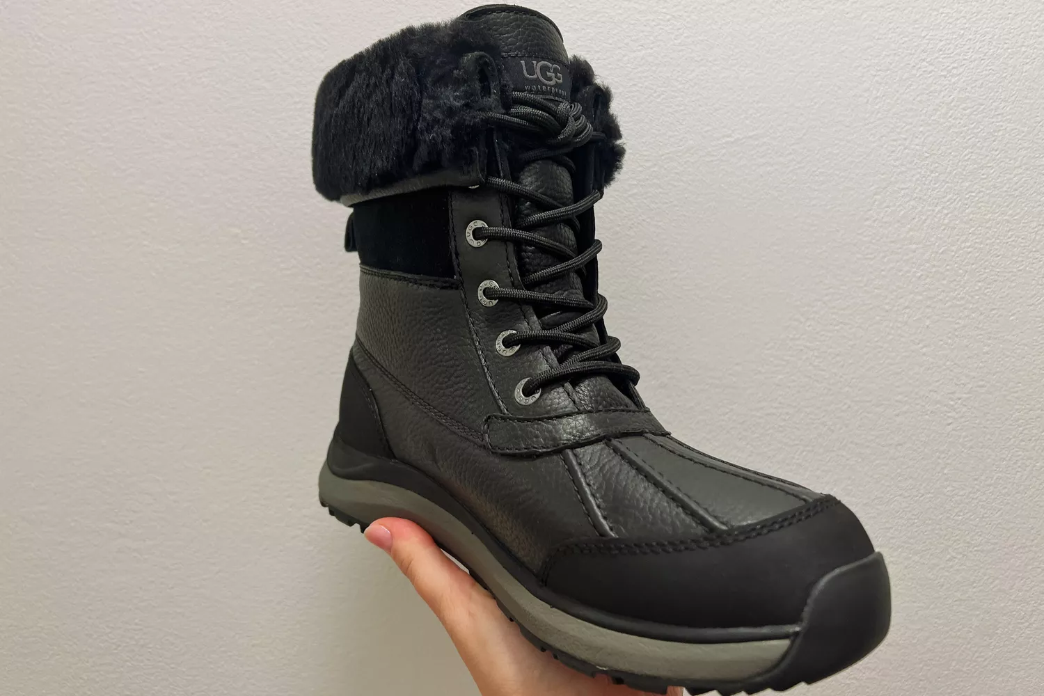 A hand holds up the UGG Adirondack III Boot in front of a gray wall