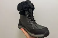 A hand holds up the UGG Adirondack III Boot in front of a gray wall